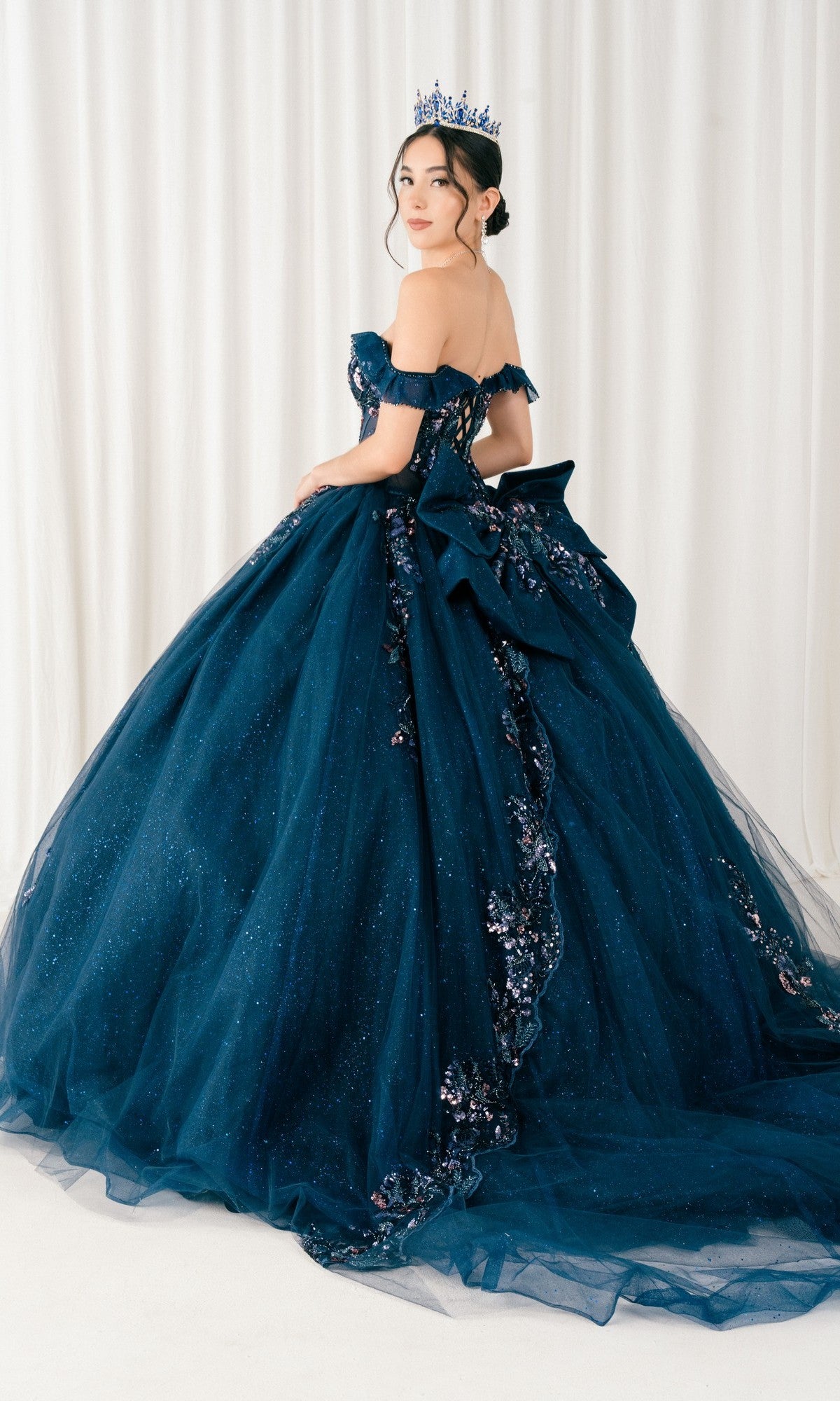 A woman in the Quinceanera Dress 8440JQ by Cinderella Couture, an off-the-shoulder navy blue ball gown with floral embellishments and a matching crown, stands in front of white curtains.